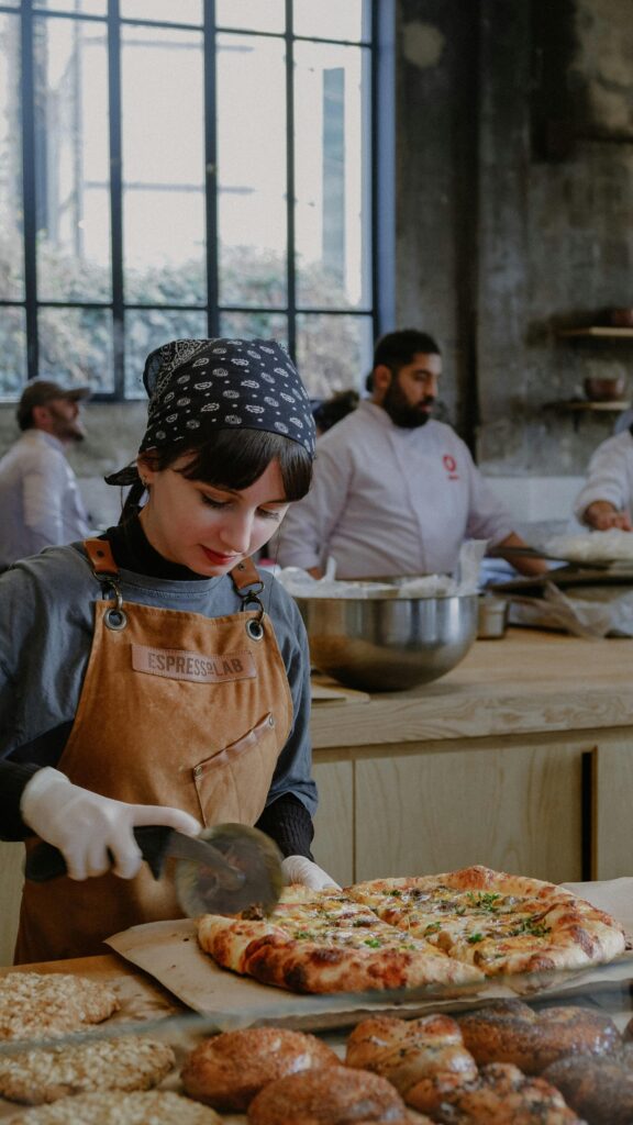 Young female chef slicing pizza in bustling bakery kitchen with breads and coworkers.